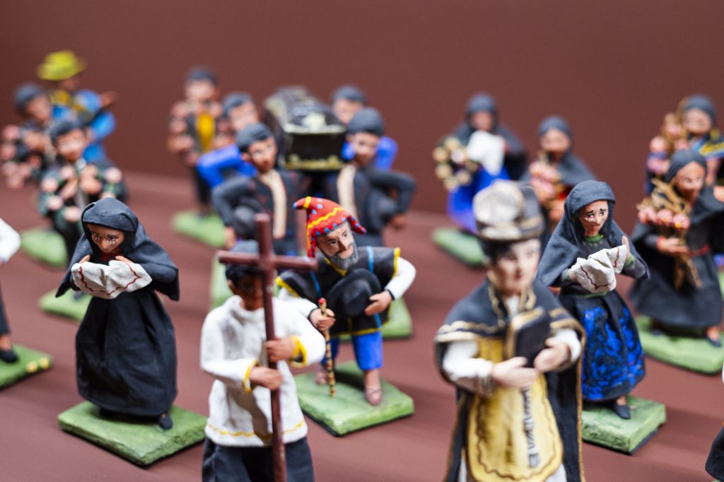 Viele kleine Keramikfiguren, die eine Begräbnisszene in Peru darstellen. Im Vordergrund ein Priester, eine Figur mit einem Holzkreuz und zwei trauernde Frauen mit schwarzen Umhängen, in der Bildmitte eine Figur mit einer bunten Ohrenkappe und dahinter Sargträger mit einem Sarg.