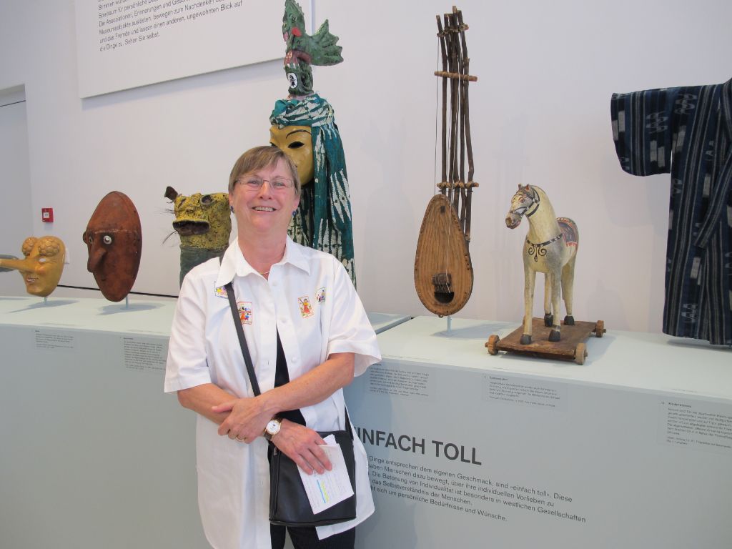 Frau Helena Roth steht in der Ausstellung neben ihrem Objekt, einer braunen Laute aus Holz. 
