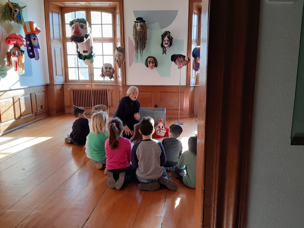 Ein Dutzend Kinder sitzen im Halbkreis in der Ausstellung zur Basler Fasnacht und hören Margrit Gontha zu.