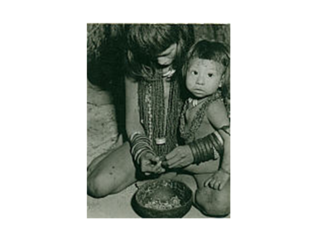Das Schwarz-Weiss-Foto zeigt eine Mutter eines indigenen Volkes. Den einen Arm hat sie um ihr Kind gelegt, da auf einem ihrer Beine sitzt. Mit beiden Händen macht sie Schmuck. Vor ihr steht eine Schüssel, gefüllt mit Schmuck.
