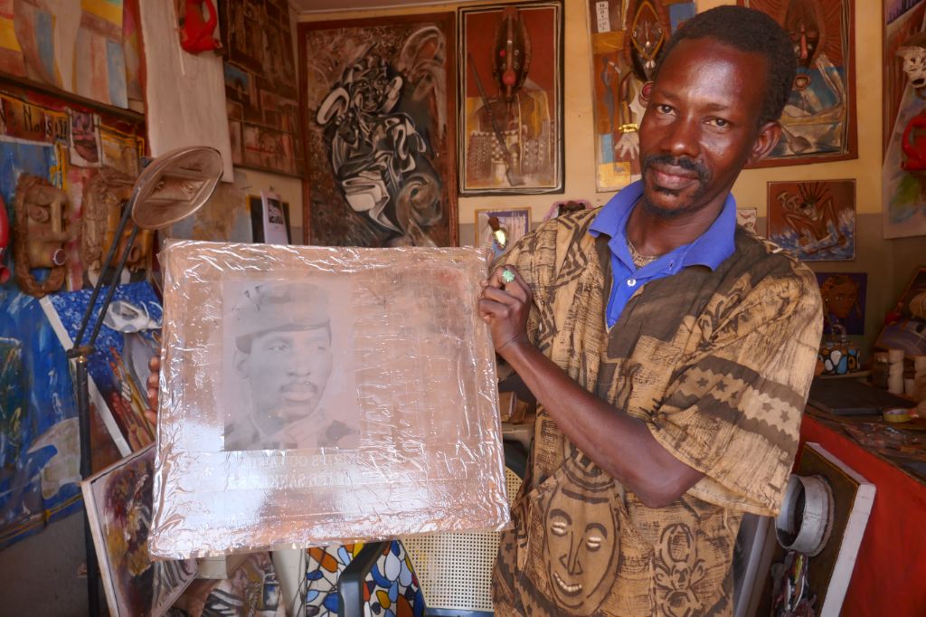 Das Foto zeigt den Künstler IB in einem Raum voller Bilder. Er hält ein Bild des Ex-Präsidenten Thomas Sankara in den Händen.
