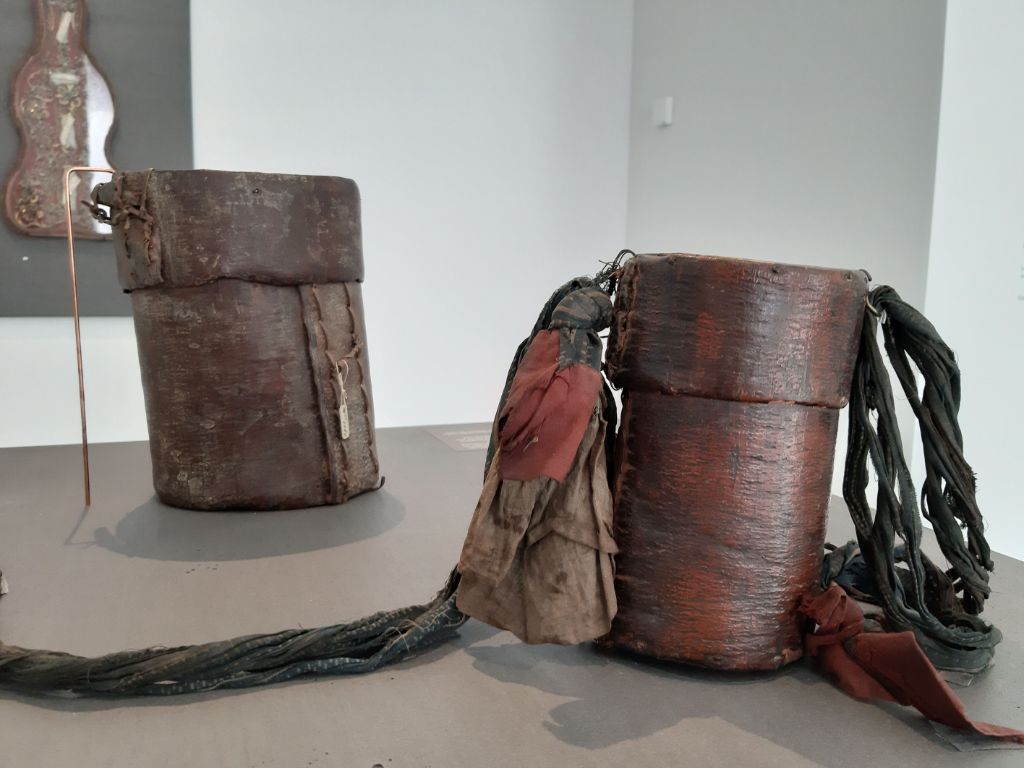 Auf grauem Podest stehen zwei rötlich-braune Lederbehälter in Röhrenform mit Deckel. Am Deckel des vorderen Behälters sind dunkle Seile und rot-braune Tücher befestigt.