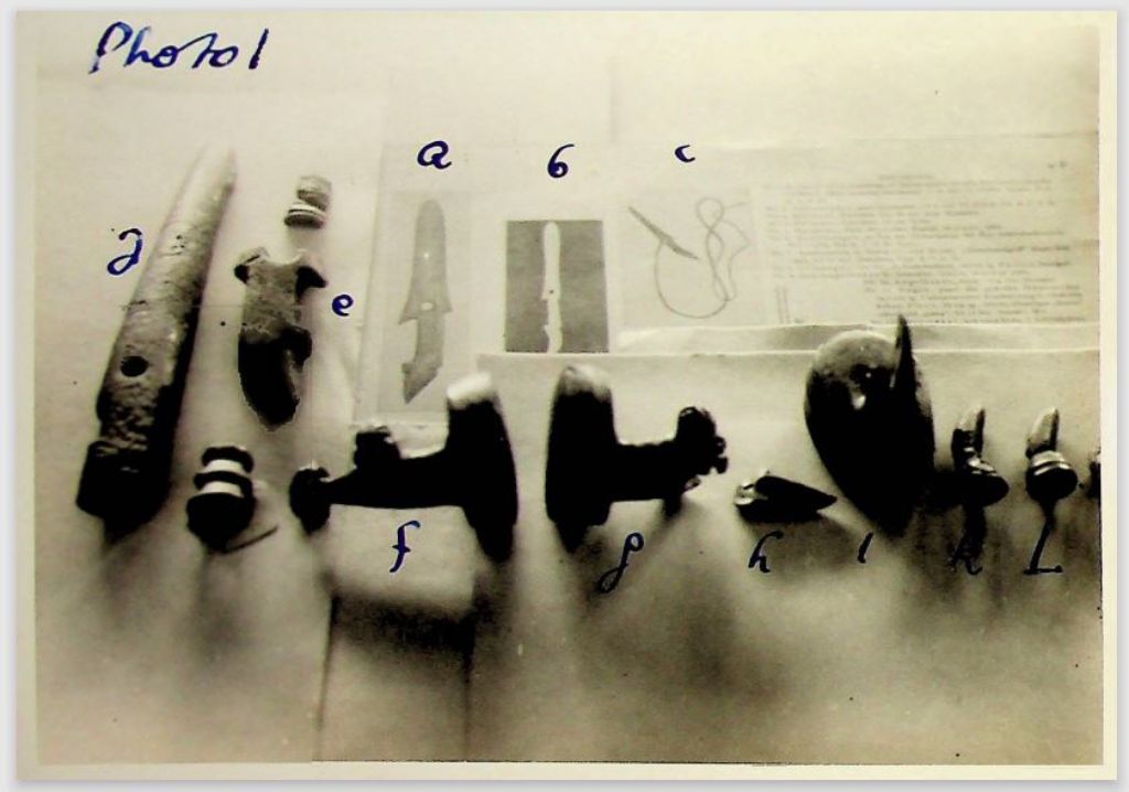 Ein Schwarz-Weiss-Foto auf dem verschiedene Gegenstände zu sehen sind auf einer Platte und dahinter Bilder und Schriftstücke. Das Foto ist mit Phot 1 angeschrieben und die Gegenstände sind nummeriert oder mit Kleinbuchstaben versehen.