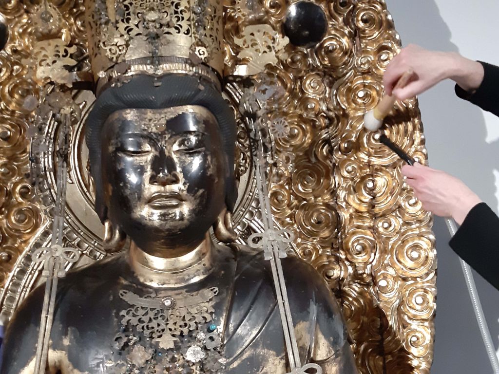 Kopf und Schultern einer schwarz-goldenen Buddhastatue mit einer goldenen Gloriole dahinter, deren Blumenmotive von zwei Händen geputzt werden. Die rechte Hand hält einen Pinsel, die linke Hand den obersten Teil eines kleinen Staubsaugers.