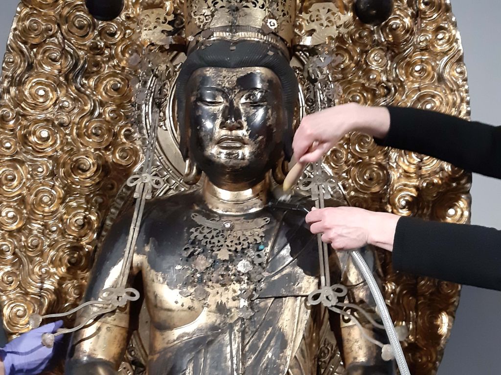Drei Hände reinigen eine golden-schwarze Buddhafigur, von der man Kopf und Oberkörper sieht. Eine Hand in blauem Handschuh hält einen Pinsel und staubt den rechten Arm ab. Zwei Hände reinigen die linke Halspartie des Buddha, mit Pinsel und Staubsaugerschlauch.