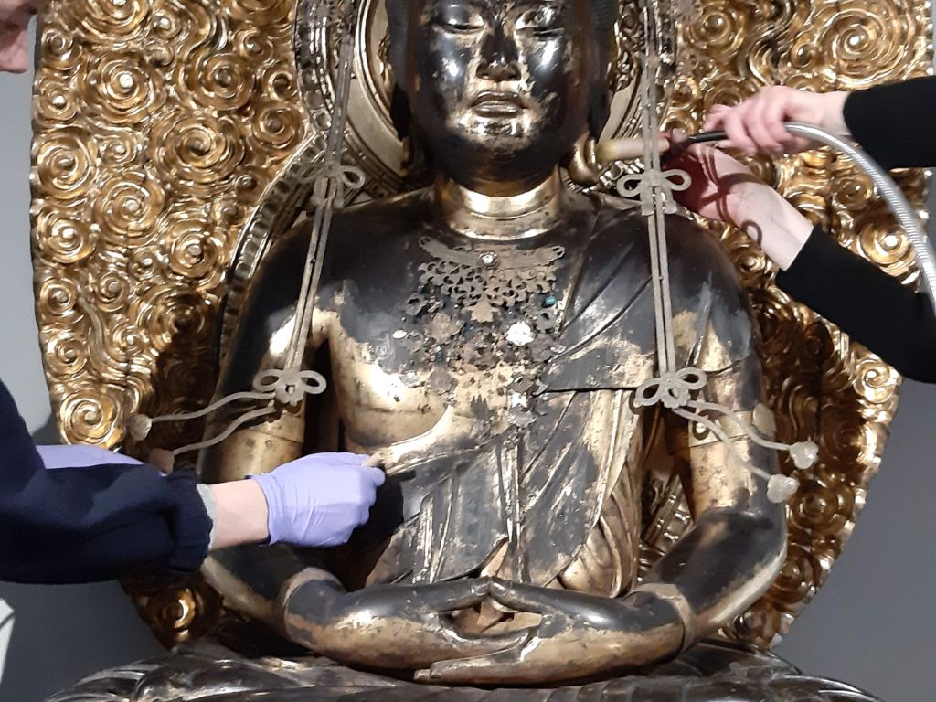 Drei Hände reinigen eine schwarz-goldene Buddhastatue, von der man Kopf und Oberkörper sieht. Eine Hand in blauem Handschuh pinselt am Kleid des Buddha in der Mitte des Körpers. Zwei weitere Hände putzen mit Pinsel und Staubsaugerschlauch den Rücken.