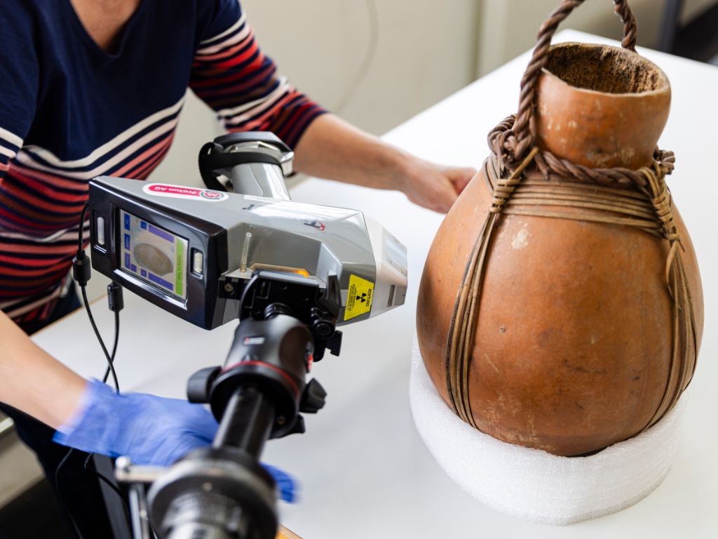 Eine Art Kamera ist auf einem Stab montiert und wird an einen braunen Krug gehalten, der mit Seilen umspannt ist und in einem weissen Gefäss sitzt.