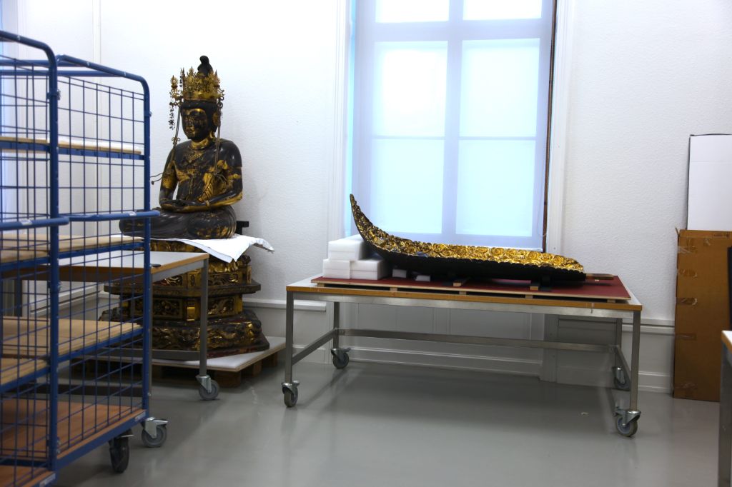 In einem Raum mit weissen Wänden sitzt in der linken hinteren Ecke ein gold-schwarzer Buddha, der menschengross ist. Vor ihm steht ein Roll- und davor ein blauer Gitterwagen. Neben ihm befindet sich ein Fenster mit undurchsichtigem weissem Rollo. Davor steht ein Rolltisch, auf dem etwas Flaches, Goldenes liegt, das links leicht nach oben geschwungen ist.