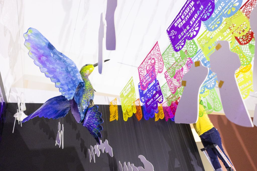 An einer weissen Decke hängen farbige Scherenschnitte in der Grösse von A3-Blättern. Ebenfalls hängen weisse Figuren von der Decke sowie eine Vogelfigur mit grünem Kopf, grüner Brust und violett-blauen Flügeln und Schwanzfedern.