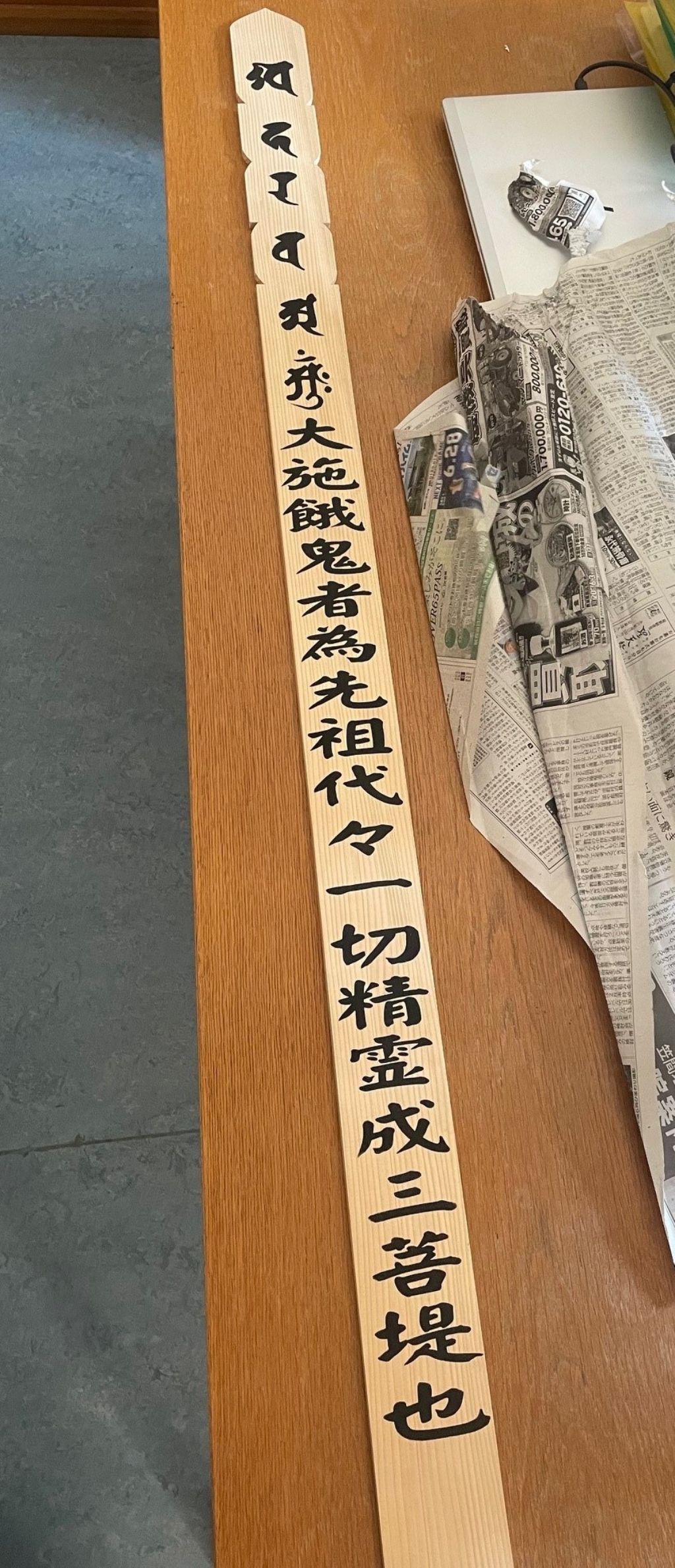 Auf einem Holztisch liegt ein langes helles Holzbrett mit Einkerbungen seitlich und schwarzen Schriftzeichen darauf gemalt. Daneben liegt eine zerknüllte Zeitung.