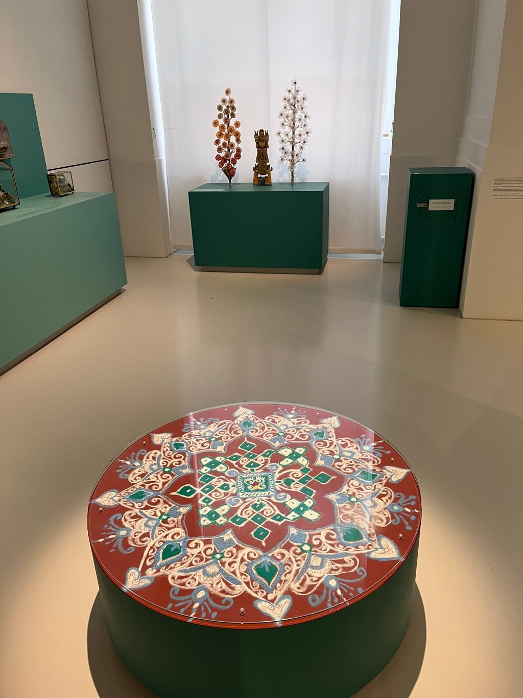 Ein rundes Tischchen, verziert mit einer plattenfüllenden Blüte in verschiedenen Farben steht in einem grossen Saal. Im Hintergrund sieht man drei Podeste, auf denen Figuren stehen. Und ein raumhohes Fenster mit weissem Vorhang.