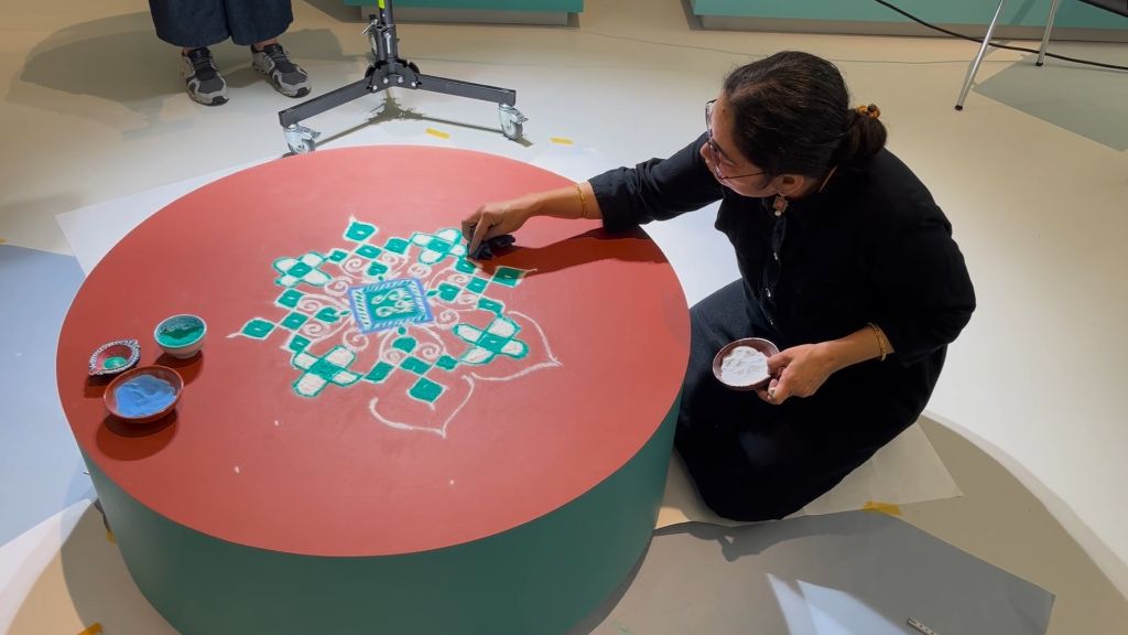 Eine Frau mit dunklem Haar, zusammengenommen in einem Rossschwanz, in dunklen Kleidern kniet vor einem runden roten Tischchen. In der linken Hand hält sich ein Schüsselchen mit weissem Pulver. Mit der rechten Hand streut sie etwas auf den Tisch. Auf der Tischplatte ist der Anfang eines Musters zu sehen, es ist eine Blüte in Weiss und Türkis.