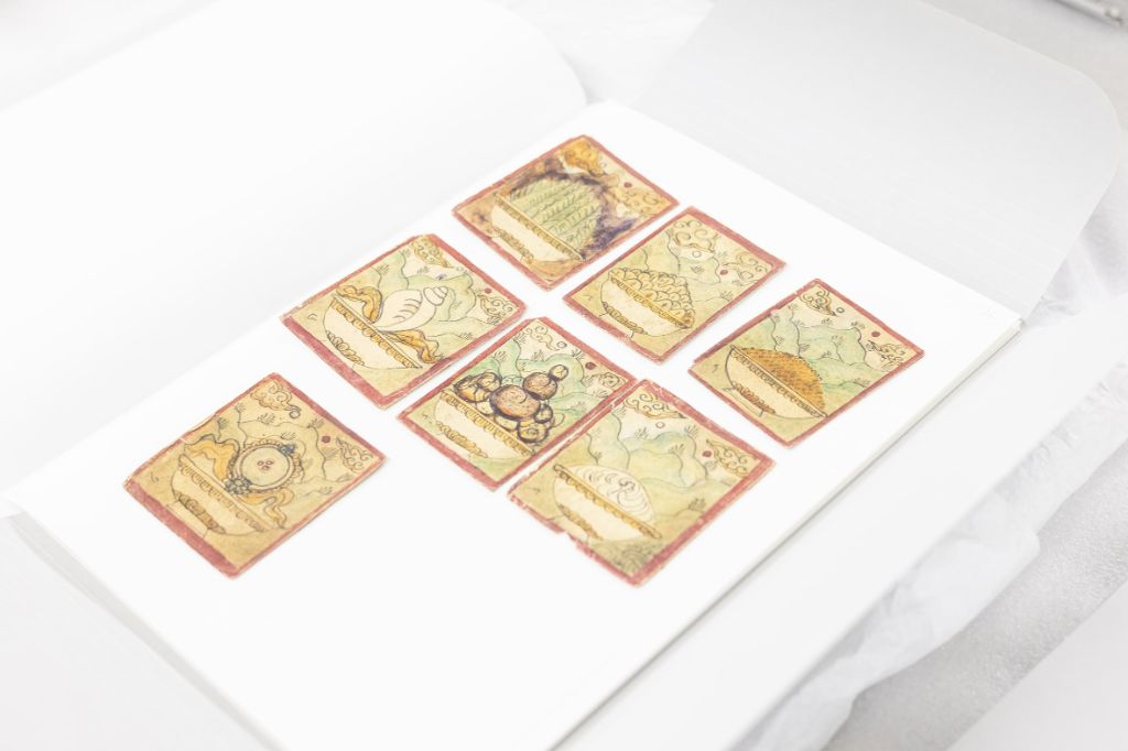 Auf einem weissen Tisch liegen sieben kleine Karten – ähnlich Spielkarten –in einem aufgeschlagenen Umschlag. Sie sind orange umrandet und zeigen verschiedene Sujets in grün-gelb-beigen Tönen. 