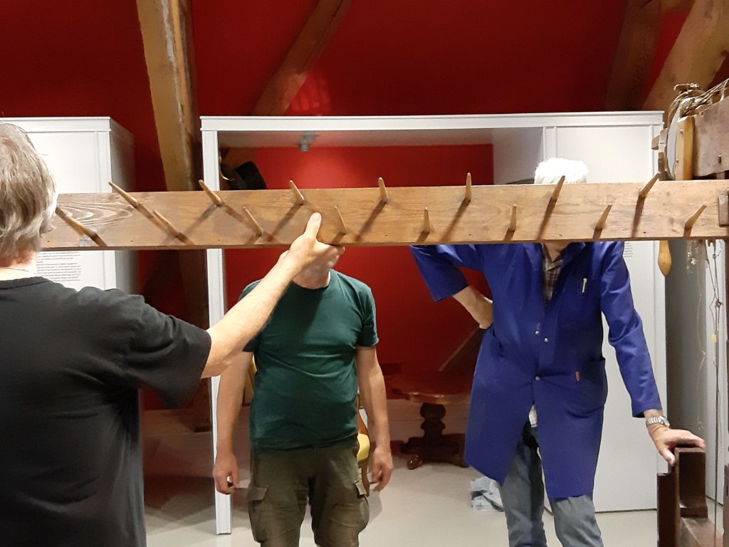 Ein Mann im schwarzen T-Shirt mit grauem Haar hält einen Balken mit vielen herausragenden runden Hölzchen in den Händen. Zwei weitere Männer stehen im Gegenüber. Ihre Gesichter sind vom Balken verdeckt.