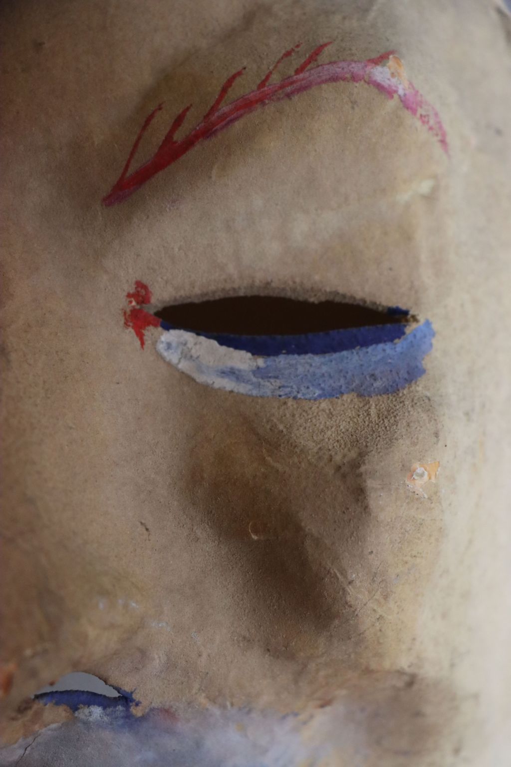Von einer Maske sieht man den Augenschlitz. Er ist weiss-hellblau untermalt. Die Augenbraue ist rot.
