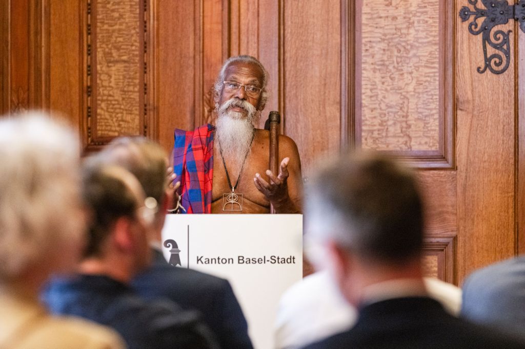An einem weissen Rednerpult, auf dem in schwarzer Schrift Kanton Basel-Stadt steht, spricht ein Mann zu einem Publikum, von dem man nur verschwommene Köpfe von hinten sieht. Der Mann hat weisses Haar und trägt einen langen weissen Bart. Er hat ein rot-blaues Tuch über der Schulter und einen markanten Anhänger um den Hals.