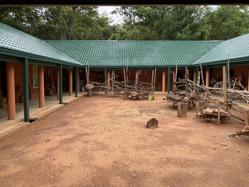 Ein einstöckiges Haus mit Veranda und grünem Ziegeldach umgibt einen rechteckigen erdigen Innenhof. Im Hof sind Teile von Bäumen aufgeschichtet.