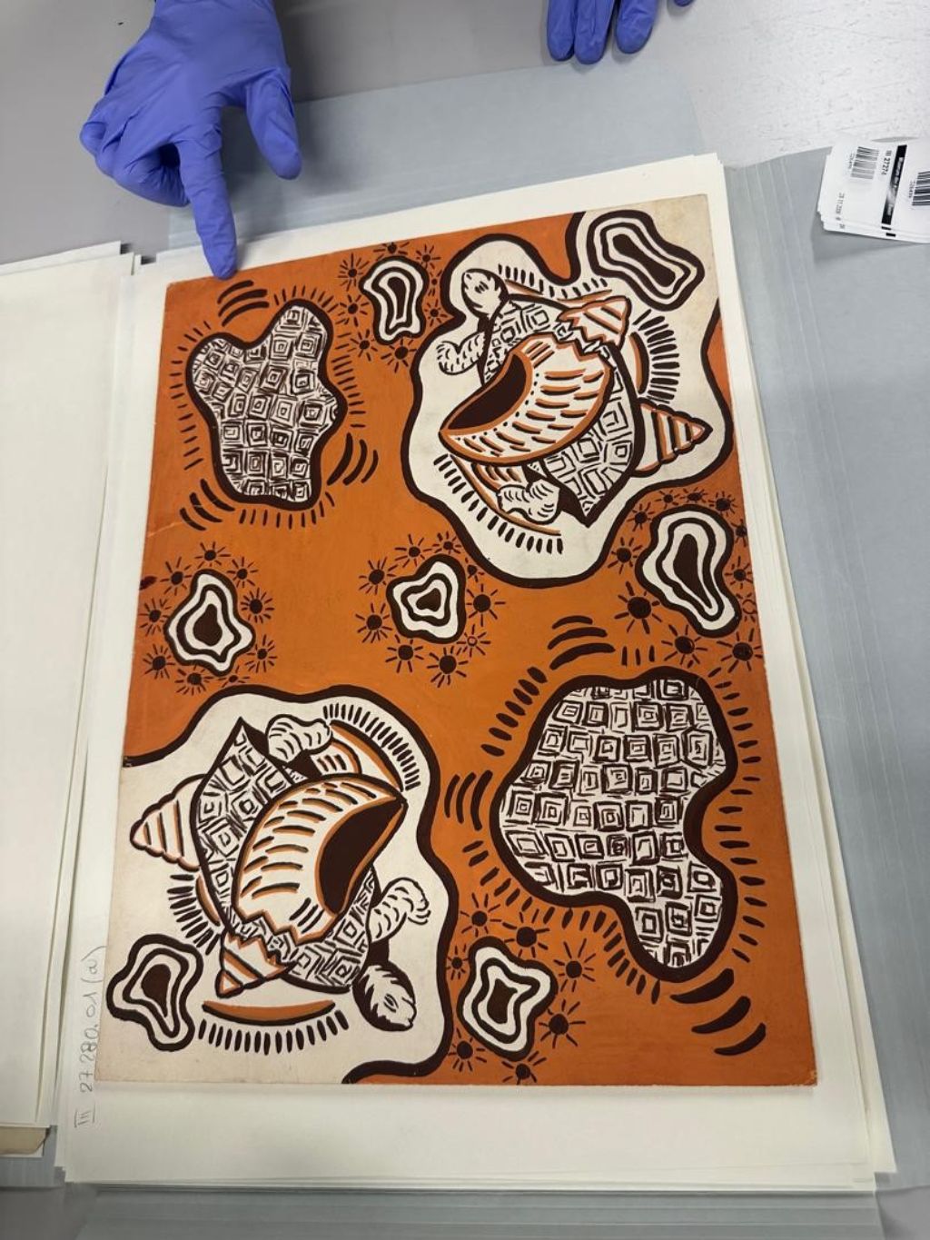 Ein rechteckiges Stück Stoff liegt auf einem Tisch. Es ist orange mit schwarz-weissen Figuren. Eine Hand in blauem Handschuh zeigt mit dem Zeigfinger auf den Stoff.