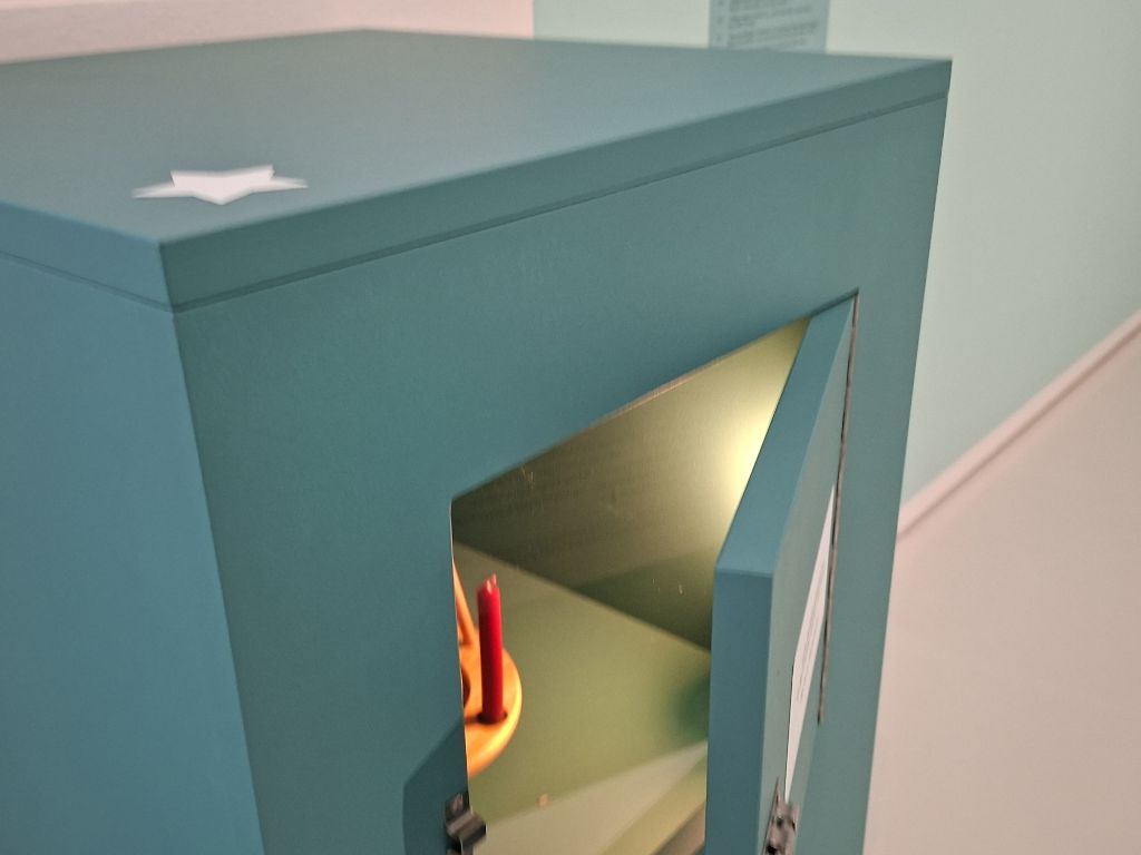 In einem türkisfarbenen viereckigen Kasten ist ein Türchen geöffnet. Im Kasten entdeckt man eine kleine rote Kerze auf einem Holzbrett. Mehr sieht man nicht. Auf dem Kasten klebt ein weisser Stern.