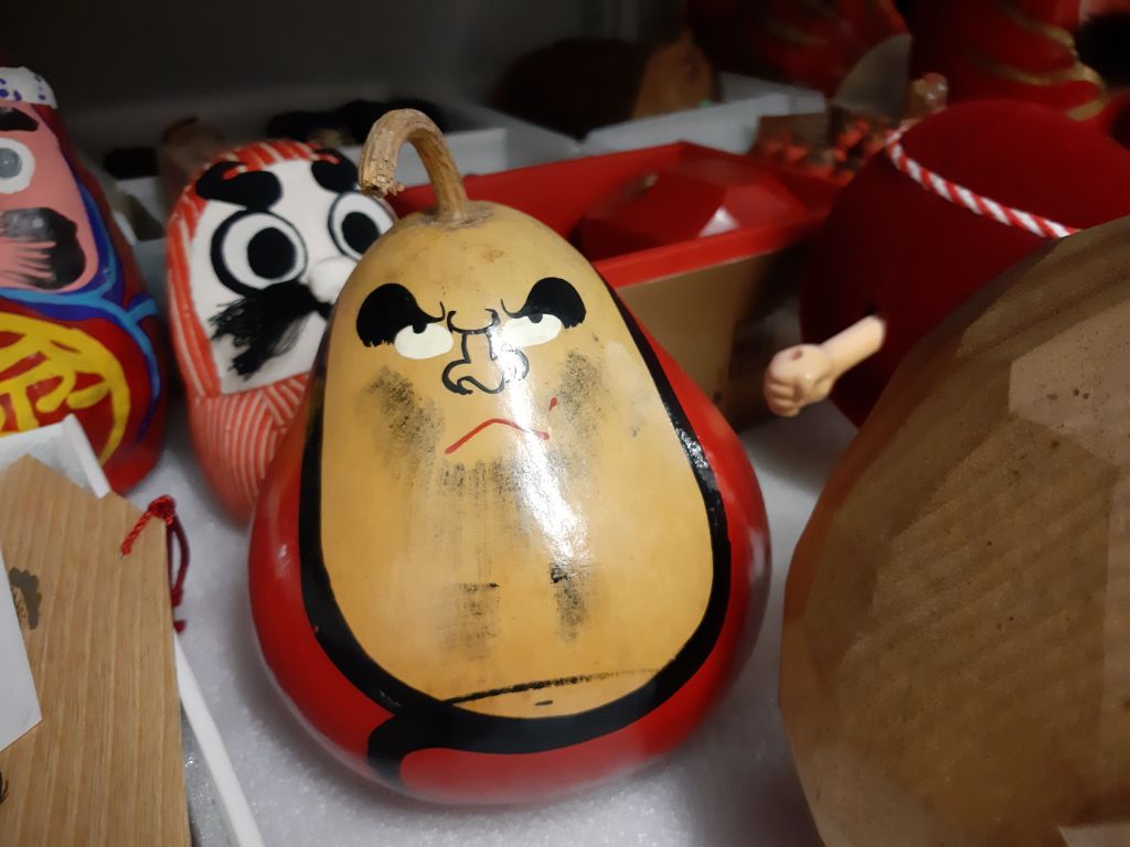 Ein birnenförmiges Objekt liegt auf einem Gestell. Es hat einen roten Körper und ein langes holzfarbenes Gesicht und schaut grimmig drein. Der Stiel wölbt sich wie ein Haarschopf oben aus der Figur. Daneben und dahinter stehen und liegen ähnliche Figuren.