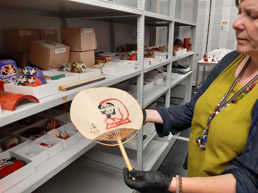 Eine Frau in olivem Kleid mit dunkelblauer Jacke und farbiger Kette um den Hals hält in den Händen einen hölzernen Fächer. Auf diesem kreisrunden Fächer ist eine kauernde Figur gezeichnet, in Rot und Schwarz. Dahinter liegen und stehen in einem Regal noch viele kleine Figuren.