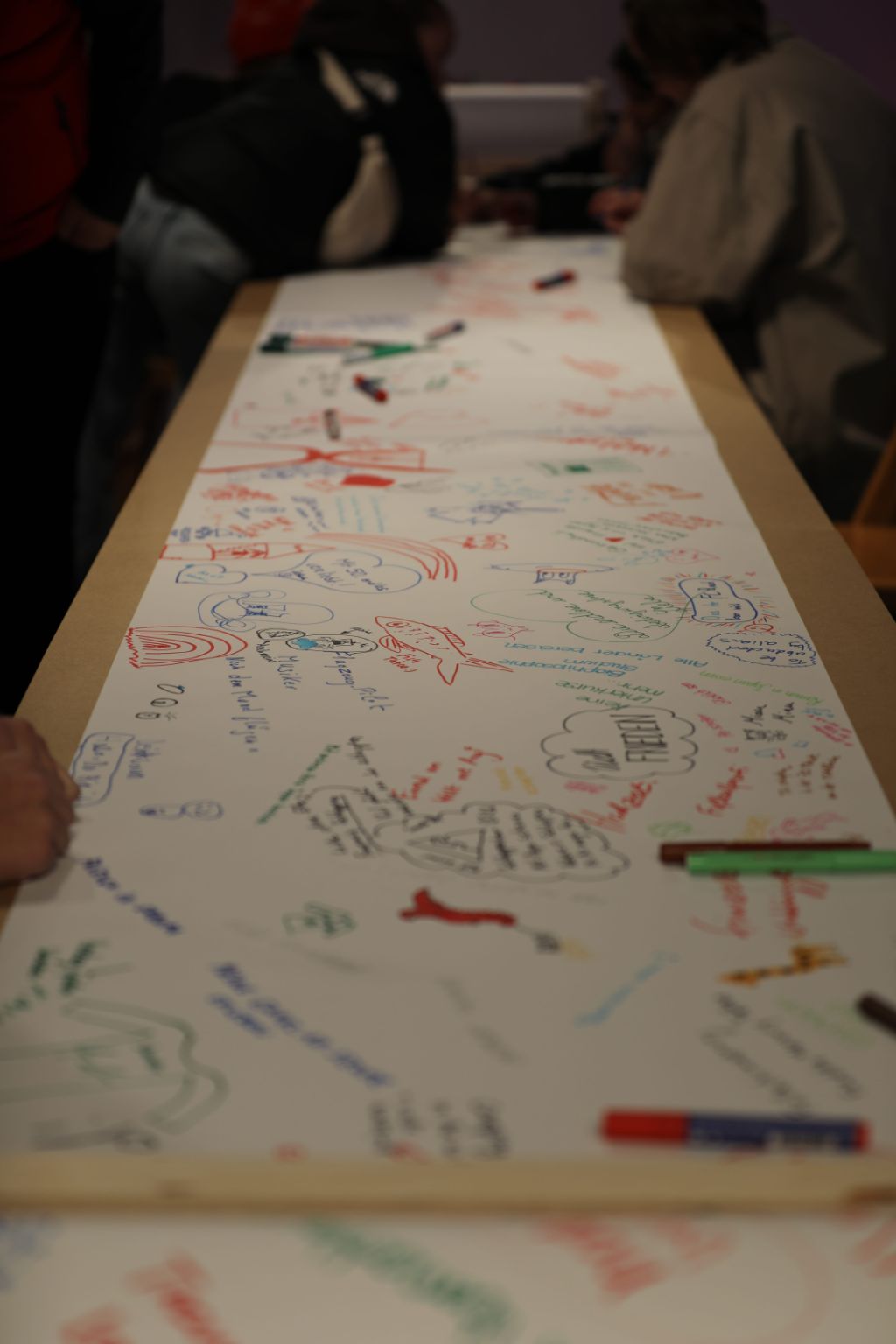 Auf einem langen Tisch ist eine weisse Papierrolle ausgerollt, auf der mit Filzstiften ganz viel geschrieben steht, kreuz und quer. Es sind auch kleine Zeichnungen zu sehen, Figuren und Berge.