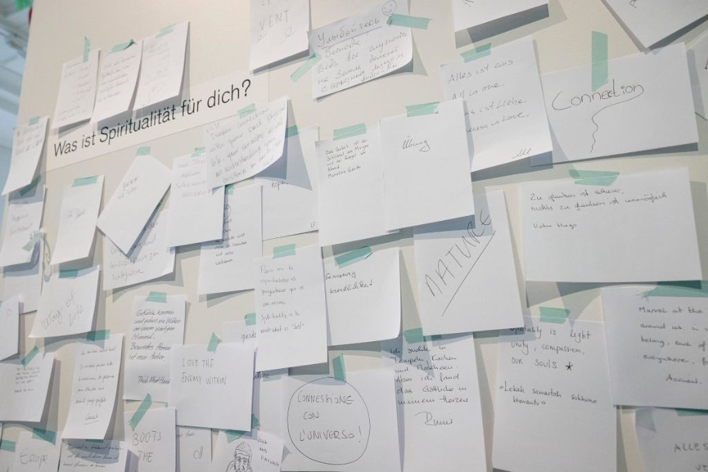 Weisse Wand, auf der viele kleine weisse Zettel mit grünen Klebern festgemacht sind. Auf den Zetteln stehen Worte und Sätze. Links oben auf der Wand steht die Frage: Was ist Spiritualität für dich?
