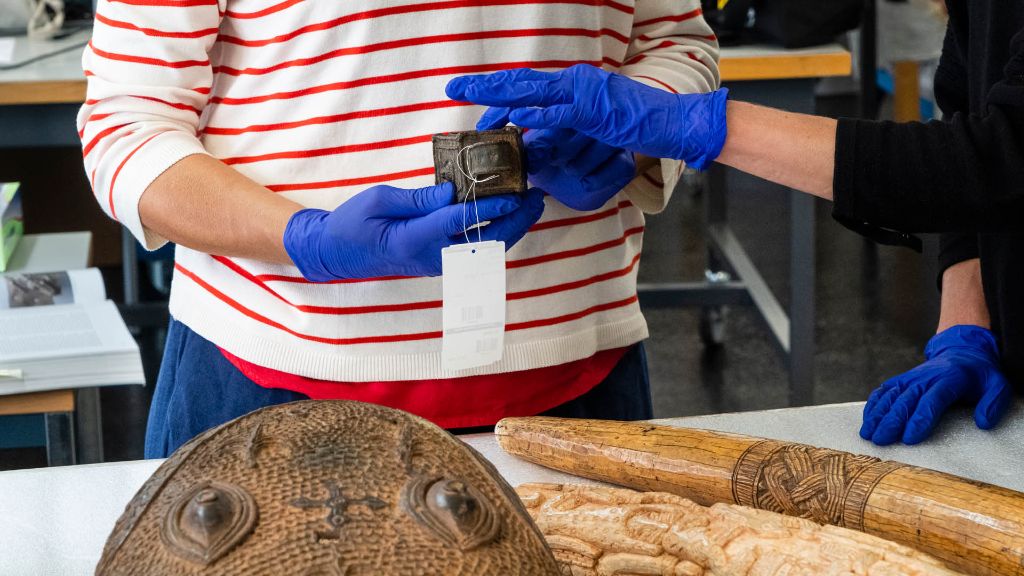 Zwei paar Hände in blauen Gummihandschuhen untersuchen Objekte aus Benin / Nigeria. Ein kleineres Objekt mit einem Etikett wird von Händen gehalten, weitere Objekte, darunter Elfenbein, liegen auf einem Tisch.