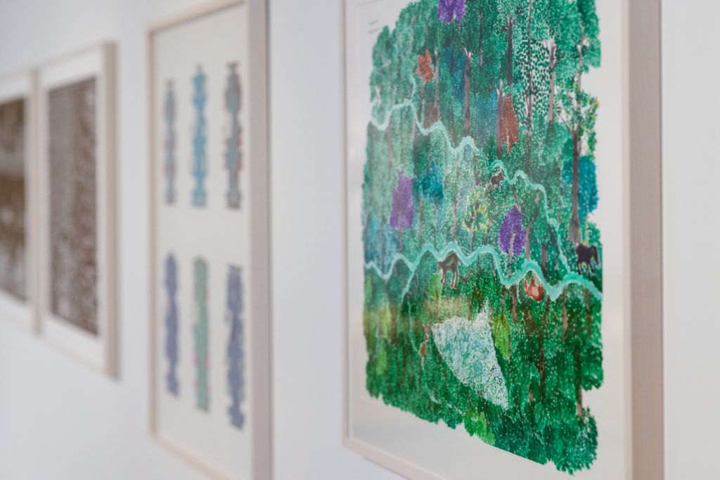 Wand mit vier weiss gerahmten Bildern in schräger Ansicht, das vorderste Bild zeigt eine grüne Waldlandschaft, zwei ineinanderlaufenden Flüssen, Tieren und bunten Pflanzen. Die anderen drei Bilder sind nur verschwommen zu erkennen.