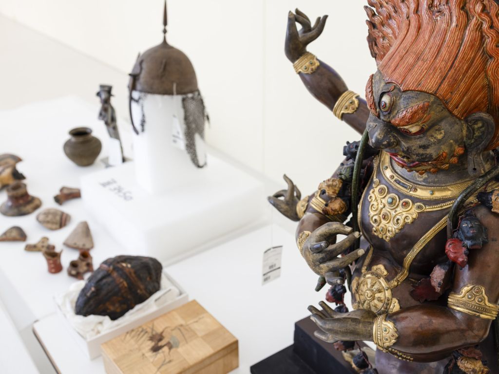 Am rechten Bildrand eine grosse Figur aus dunklem Holz mit mehreren Armen, goldenen und roten Verzierungen. Links davon auf einem Tisch mehrere kleine Objekte aus Holz und Metall, zu erkennen sind eine Maske sowie eine metallene Kopfbedeckung