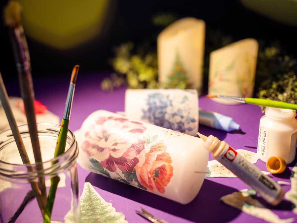 Mit Blumen und weihnachtlichen Mustern verzierte Kerzen liegen und stehen auf einem violetten Hintergrund. Dazu geöffnete Farbtöpfe, Pinsel und Wachsfarbe aus der Tube.