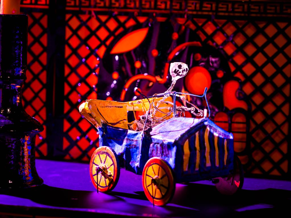 Mexikanische Totenfigur aus Pappmache, in Form eines Skeletts, welches ein blaues Oldtimer-Auto mit offenem gelben Verdeck, roten Rädern mit gelben Felgen steuert. Der Boden ist blau, im Hintergrund eine rot beleuchtet Wand mit einem Scherenschnitt eines Totenkopfs