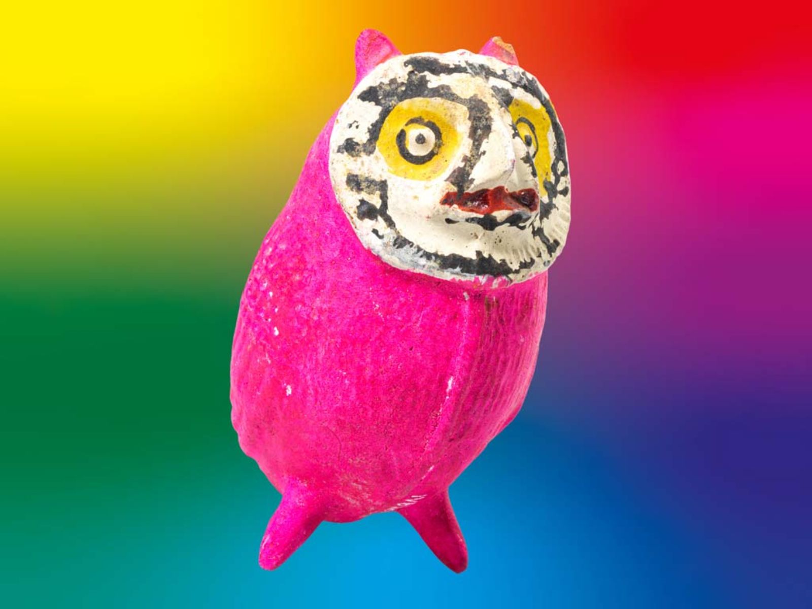Eulenfigur mit weissem Gesicht, gelb umrahmten Augen und knallig pinkem Körper, vor einem knallbunten, regenbogenfarbigen Hintergrund